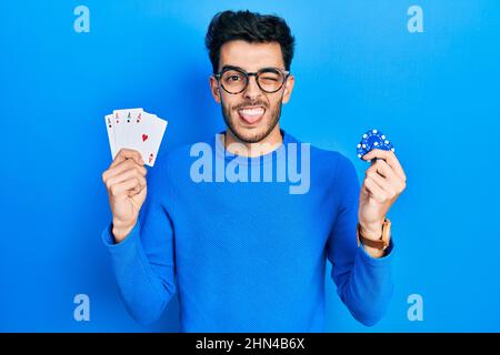 Jeune homme hispanique jouant au poker tenant des jetons de casino et des cartes collant la langue dehors heureux avec l'expression drôle. Banque D'Images