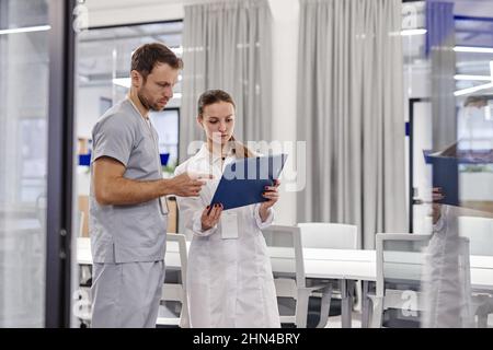 Jeune femme généraliste en blouse de laboratoire montrant un document médical à un collègue tout en le consultant au sujet de l'information Banque D'Images