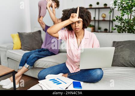 Mère de famille interraciale travaillant à l'aide d'un ordinateur portable à la maison rendant amusant des personnes avec les doigts sur le front faisant le mocking de geste perdant et inssul Banque D'Images