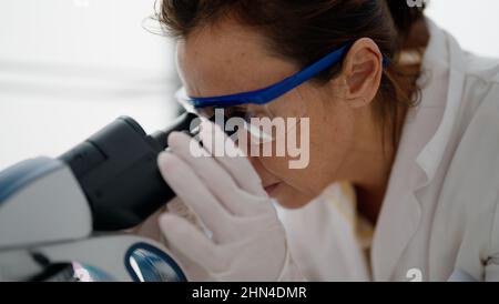 Femme hispanique d'âge moyen portant l'uniforme scientifique au microscope en laboratoire Banque D'Images