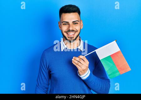 Jeune homme hispanique avec barbe portant le drapeau de madagascar qui a l'air positif et heureux debout et souriant avec un sourire confiant montrant des dents Banque D'Images