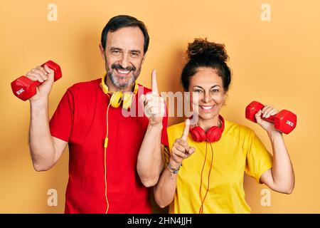 Couple d'âge moyen de femme hispanique et d'homme portant des vêtements de sport en utilisant des haltères souriant avec une idée ou une question pointant du doigt avec le visage heureux, numbe Banque D'Images