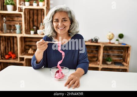 Femme d'âge moyen à cheveux gris mangeant un mètre-ruban assis sur la table à la maison. Banque D'Images