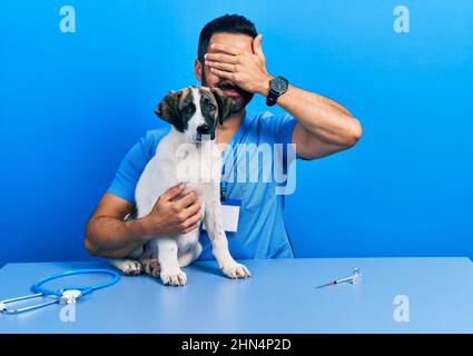 Beau vétérinaire hispanique avec la barbe de vérifier la santé de chien couvrant les yeux avec la main, regardant grave et triste. sightless, cachette et rejet conc Banque D'Images