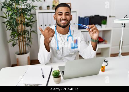 Jeune médecin indien tenant le thermomètre pointant le pouce vers le côté souriant heureux avec la bouche ouverte Banque D'Images
