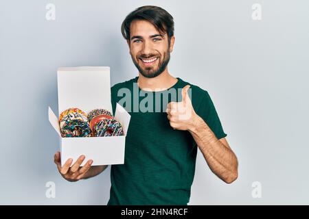 Jeune homme hispanique tenant une boîte de beignets colorés savoureux souriant heureux et positif, pouce vers le haut faisant l'excellent et signe d'approbation Banque D'Images