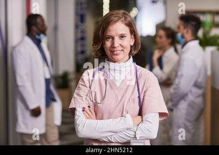 Jeune femme heureuse généraliste ou infirmière en soins médicaux se frotte croisant les bras par la poitrine tout en se tenant contre des collègues interculturels Banque D'Images