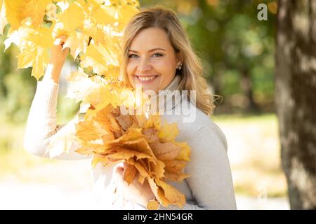 Belle femme d'âge moyen souriante aux cheveux fins ondulés dans un chandail à col roulé tenant un bouquet de feuilles d'érable et une branche. Banque D'Images