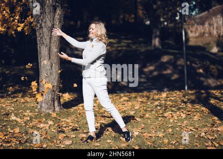 Belle-belle souriante d'âge moyen avec de longs cheveux ondulés justes dans le chandail gris à col roulé jeter les feuilles d'érable jaune. Banque D'Images