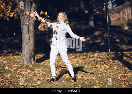 Mignonne souriante femme d'âge moyen avec de longs cheveux ondulés équitables en chandail gris à col roulé jeter une pile de feuilles d'érable jaune. Banque D'Images