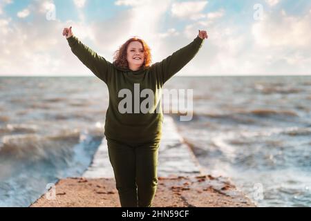 Une femme heureuse en surpoids a jeté les mains, debout sur la jetée au bord de la mer. Ciel et océan en arrière-plan. Le concept du succès. Banque D'Images