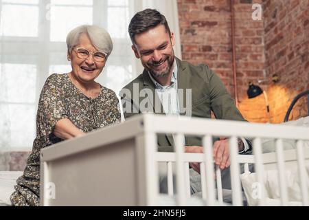un homme d'âge moyen caucasien et sa mère aux cheveux gris souriant au nouveau-né dans un berceau. Photo de haute qualité Banque D'Images