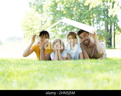 Concept d'hypothèque et de logement pour les jeunes familles. Mère père et deux enfants allongé sur l'herbe et portant le symbole du toit Banque D'Images