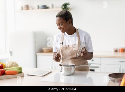Bonne jeune femme afro-américaine dans la cuisine de tablier et regarder la leçon vidéo sur tablette Banque D'Images
