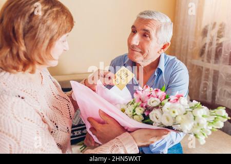 Bonne journée pour les femmes, cadeau du 8 mars. Homme senior doutant son épouse bouquet de fleurs à la maison. Surprise heureuse femme lisant la carte de voeux. Couple familial cel Banque D'Images