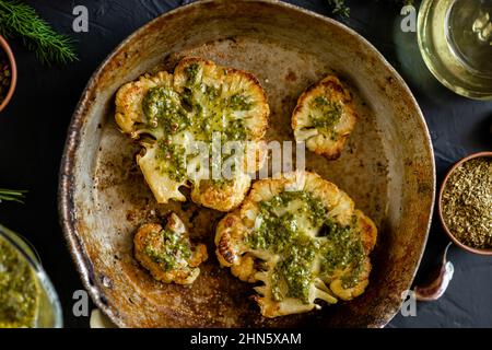 Le steak de chou-fleur aux épices se trouve dans une poêle. Huile d'olive, sauce chimichurri, herbes, diverses épices côte à côte. Arrière-plan sombre. Plats végétariens Banque D'Images