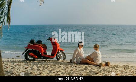 Trajet en scooter. Un couple charmant sur une moto rouge vêque de vêtements blancs sur une plage de sable. Les gens juste mariés embrassent les hugs marchant près des palmiers tropicaux, la mer. Lune de miel de mariage au bord de l'océan. Location de motos. Banque D'Images