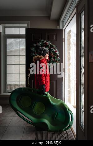 Garçon en manteau rouge et chapeau debout dans la porte avec traîneau le jour d'hiver. Banque D'Images