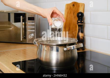 Vue rapprochée de la main de la femme versant du sel à l'eau bouillante sur la cuisinière électrique. Faire passer le sel sur le pot à plat en acier inoxydable. Banque D'Images