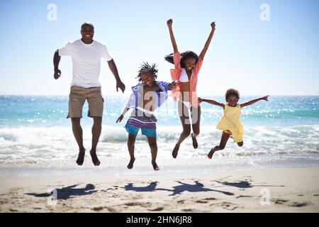 Woohoo Vacation Time Rocks. Une famille afro-américaine excitée sautant dans les airs sur la plage avec un bel océan en arrière-plan. Banque D'Images
