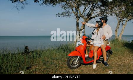 J'adore un couple sur une moto rouge vêque de vêtements blancs sur une route forestière. Deux caucasiens de touristes femme et homme baiser des hugs assis sur le scooter. Location de moto casque de sécurité lunettes de soleil. Asie Thaïlande tourisme. Banque D'Images