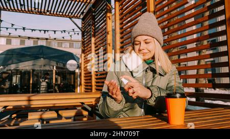 Une jeune femme dans une veste chaude traite ses mains avec un antiseptique à l'extérieur. Elle est située dans un espace urbain élégant avec un décor en bois. Banque D'Images