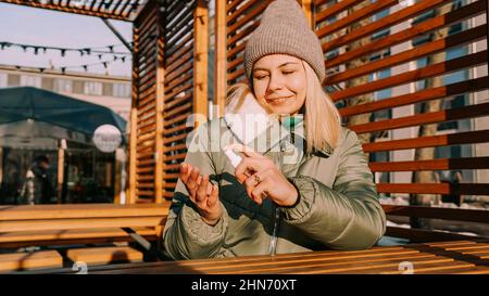 Une jeune femme dans une veste chaude traite ses mains avec un antiseptique à l'extérieur. Elle est située dans un espace urbain élégant avec un décor en bois. Banque D'Images