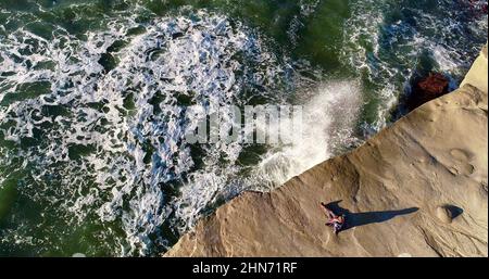 Vue aérienne d'une femme assise au bord de la falaise, regardant le coucher du soleil au parc naturel de Sunset Cliffs pendant que les vagues se écrasont sur des rochers, San Diego, Californie, États-Unis Banque D'Images