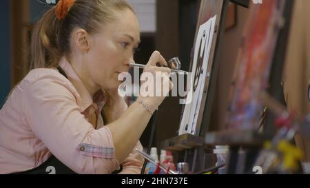 Une artiste apprend à peindre avec un aérographe avec du colorant acrylique, du papier et du chevalet. Intérieur. Concept art moderne, aérographe, aérographie, dessin pulvérisation image créer pulvérisateur dessin graffiti aérosol spray Banque D'Images
