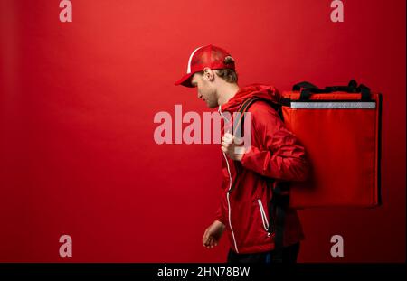 Vue latérale d'un jeune coursier professionnel dans un uniforme rouge et avec un sac thermique sur une épaule sur fond rouge couleur, espace pour le texte. Livraison de nourriture Banque D'Images