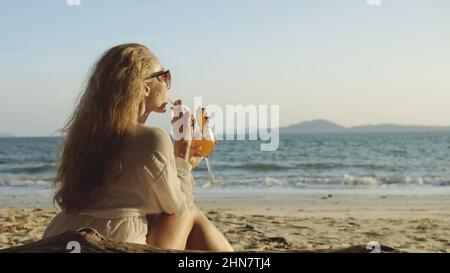 Belle femme assise dans une robe blanche lunettes de soleil, boire un cocktail d'ananas Pina Colada. Belle plage, vagues au coucher du soleil. Concept Rest tropical Resort voyager tourisme heureux vacances d'été Banque D'Images