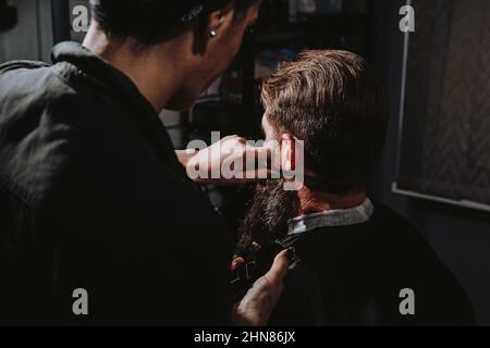 Femmes salon de coiffure professionnel servant client avec une grosse barbe épaisse par tondeuse, beau brutal jeune homme élégant obtenir une coupe de cheveux tendance, salon de coiffure noir Banque D'Images