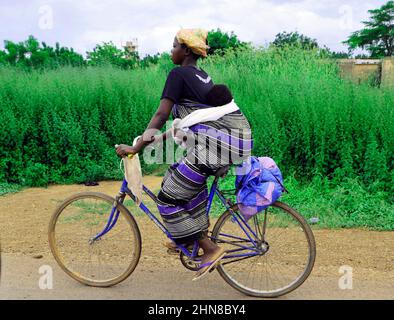 Une femme burkinabée portant son bébé sur le dos tout en montant son vélo dans le centre du Burkina Faos. Banque D'Images