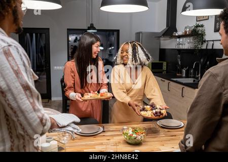 Les jeunes femmes heureux tenant des assiettes avec des hors-d'œuvre et les mettant sur la table de salle à manger, ils servent le dîner pour leurs hommes pendant la fête de dîner Banque D'Images