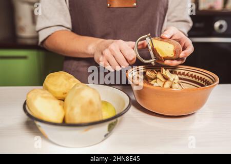 les mains de la cuisson couper le pelage des pommes de terre crues avec un couteau de cuisine pour éplucher les légumes, éplucher, mettre les déchets alimentaires dans un récipient séparé pour le traitement ultérieur. Banque D'Images