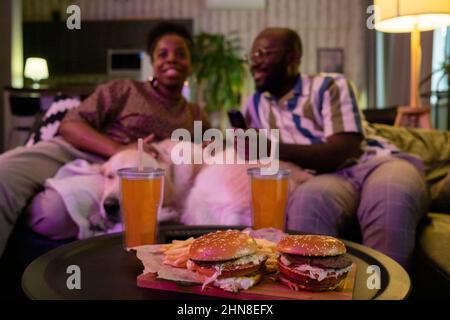 Gros plan de hamburgers et de soda sur la table avec un couple africain regardant la télévision ensemble sur le canapé en arrière-plan Banque D'Images