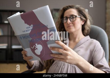 Jeune femme enceinte assise sur une chaise et lisant un livre sur la grossesse pendant qu'elle se repose après le travail au bureau Banque D'Images