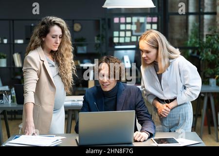 Les gens d'affaires regardant le moniteur de l'ordinateur portable et travaillant sur la présentation en ligne en équipe avec femme d'affaires enceinte qui se standign et de regarder le processus Banque D'Images