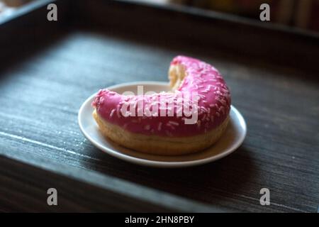 Vue latérale à moitié mangé de beignet rose vif sur une assiette ronde blanche. Arrière-plan en bois vieilli. Objet isolé. Bonbons traditionnels américains pour le petit-déjeuner Banque D'Images