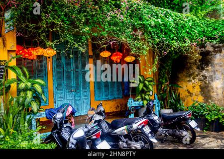 Une journée humide dans la vieille ville avec des motos garées en face d'un bâtiment jaune avec des fenêtres à volets turquoise et des vignes et des lanternes verdoyantes. Banque D'Images