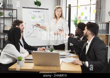 Une femme d'âge caucasien se tenant près du tableau de conférence alors qu'elle dirige une conférence pour des collègues multiraciaux. Homme et femme employé de l'entreprise en tenue officielle assis au bureau et à l'écoute du chef d'équipe Banque D'Images