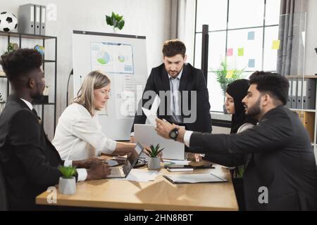 Groupe de personnes multiraciales occupées vêlées ayant discuté activement pendant la réunion au bureau. Hommes et femmes assis au bureau, utilisant des gadgets modernes et examinant divers documents. Banque D'Images