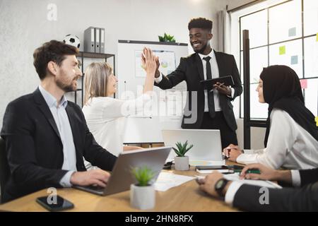 Un homme d'affaires africain positif donne cinq hauts à ses collègues multiraciaux lors du briefing au bureau. Négociation réussie de partenaires masculins et féminins. Banque D'Images