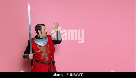 Portrait d'un guerrier médiéval choqué ou d'un chevalier portant des vêtements d'armure tenant une grande épée isolée sur fond rose. Comparaison des époques Banque D'Images
