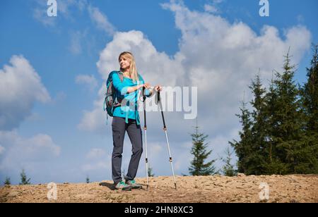 Pleine longueur de jeune femme voyageur regardant de côté et souriant tout en tenant des bâtons de trekking. Sac à dos pour randonneurs féminins debout sur une colline avec un magnifique ciel bleu sur fond. Banque D'Images