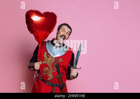 Portrait en demi-longueur d'un homme heureux et excité, guerrier médiéval en couverture protectrice avec ballon en forme de coeur rouge isolé sur fond rose Banque D'Images