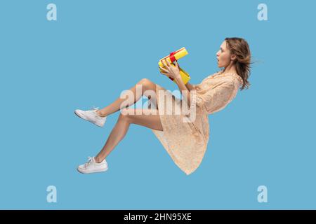 Plantant dans l'air. Femme choquée dans la robe jaune lévitant et déballant sa boîte cadeau, surpris ou choqué et regardant son cadeau d'anniversaire ou de noël. Photo d'intérieur isolée sur fond bleu. Banque D'Images