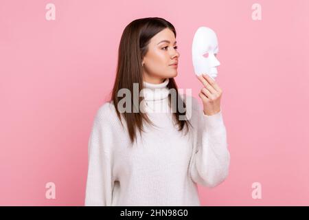 Vue latérale portrait de fille tient le masque blanc, a l'expression mystérieuse, cachant sa personnalité, portant blanc décontracté style pull. Studio d'intérieur isolé sur fond rose. Banque D'Images