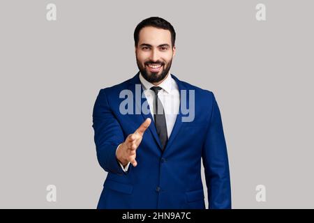 Homme barbu positif donnant la main à la poignée de main et souriant amical, consultant ou directeur des ressources humaines offrant un emploi, un partenariat, portant un costume de style officiel. Prise de vue en studio isolée sur fond gris. Banque D'Images