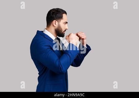Vue latérale d'un homme barbu agressif tenant les poings serrés prêts à la boxe, défendant ses droits et sa liberté, portant un costume de style officiel. Prise de vue en studio isolée sur fond gris. Banque D'Images